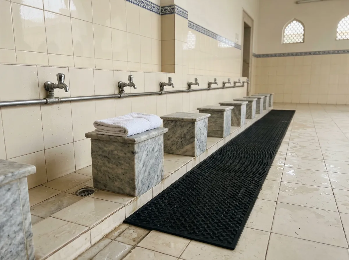 A clean tiled wudu area with a row of low marble seats in front of taps, a rubber anti slip mat along the floor and a folded dry towel placed on one seat, photographed at a low angle in daylight. No people. Photograph taken in Karachi, Pakistan. Any person shown is Pakistani and well dressed, with the face out of frame. Natural daylight, realistic colours, documentary style. No text, no signage, no logos, no watermarks. 4:3 aspect ratio.