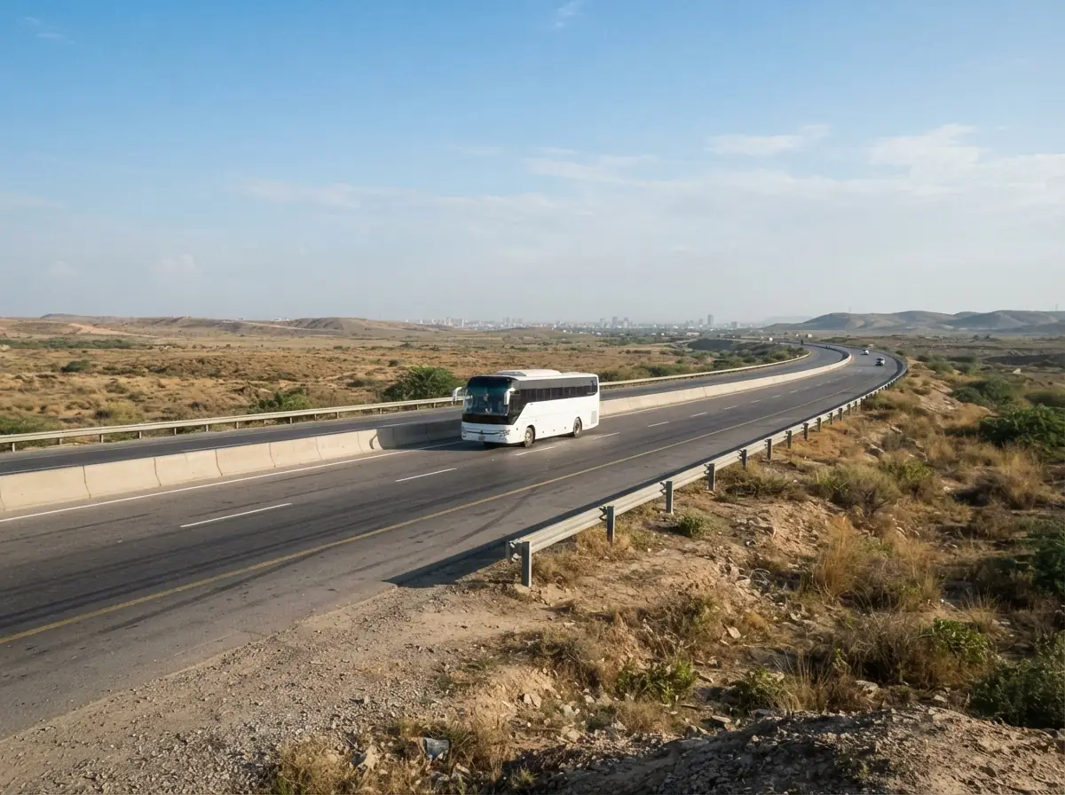 A wide view of the motorway leaving Karachi toward Hyderabad in clear morning light, with a white intercity coach in the middle distance and dry open country on both sides. No people. Photograph taken in Karachi, Pakistan. Any person shown is Pakistani and well dressed, with the face out of frame. Natural daylight, realistic colours, documentary style. No text, no signage, no logos, no watermarks. 4:3 aspect ratio.