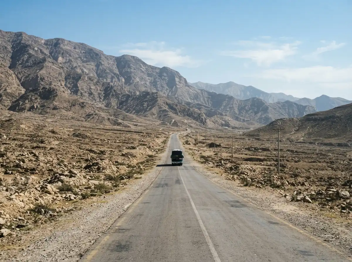 A long straight highway crossing dry rocky hills in Balochistan under a clear sky, with a single coach travelling toward the camera in the distance. No people. Photograph taken in Karachi, Pakistan. Any person shown is Pakistani and well dressed, with the face out of frame. Natural daylight, realistic colours, documentary style. No text, no signage, no logos, no watermarks. 4:3 aspect ratio.